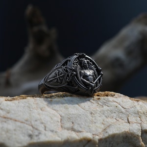 Puede incluir: Un anillo de plata detallado con un dise&ntilde;o intrincado. El anillo presenta una figura central alada que sostiene una espada y un escudo, con las palabras "SAINT MICHAEL". El anillo est&aacute; sobre un fondo neutro.