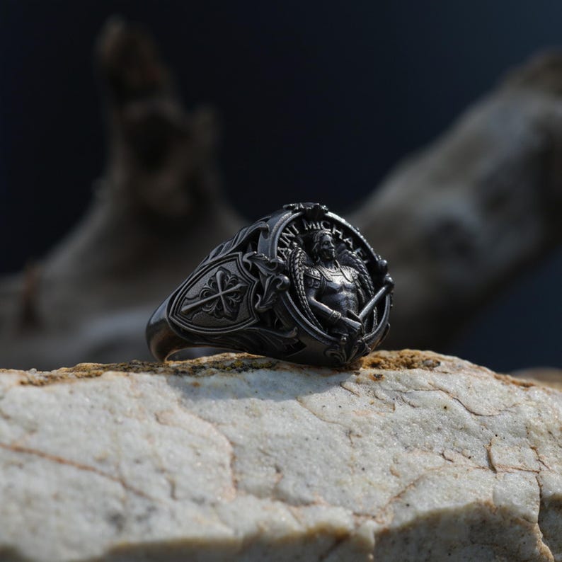 Puede incluir: Un anillo de plata detallado con un dise&ntilde;o intrincado. El anillo presenta una figura central alada que sostiene una espada y un escudo, con las palabras "SAINT MICHAEL". El anillo est&aacute; sobre un fondo neutro.