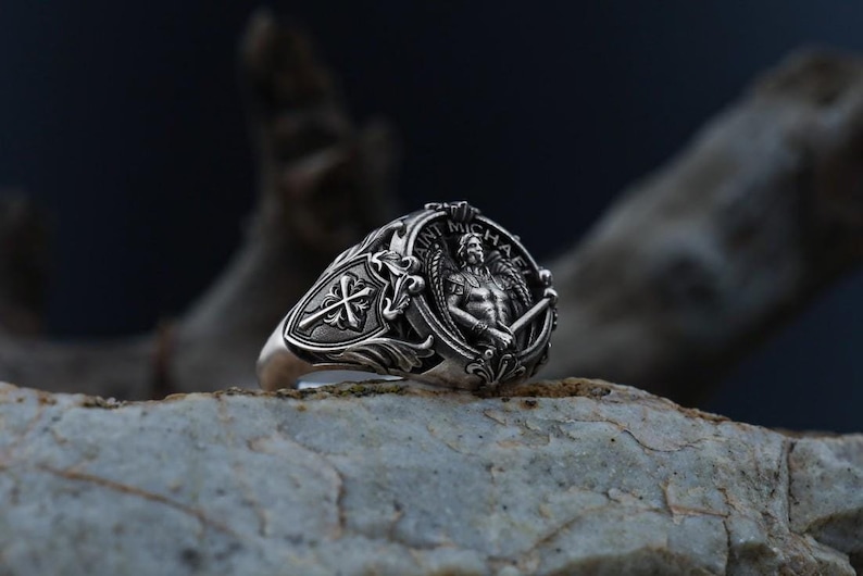 Puede incluir: Un anillo de plata con una detallada representaci&oacute;n de San Miguel con una espada, rodeado por las palabras "SAINT MICHAEL". El anillo tiene un dise&ntilde;o ornamentado con una cruz y otros elementos decorativos. El anillo se muestra sobre una superficie de piedra texturizada.