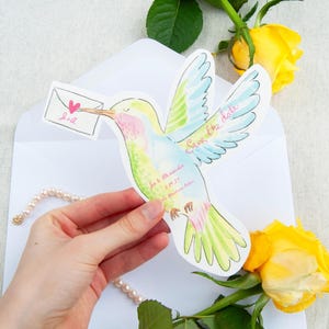 May include: A watercolour hummingbird-shaped save-the-date card with a small envelope held in the bird's beak. The card features pastel colours, the text "Save the date," and wedding details. Yellow roses and a pearl necklace are also in the image.