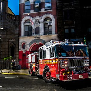 Puede incluir: Un camión de bomberos rojo y blanco con rayas amarillas está estacionado frente a una estación de bomberos de ladrillo. El camión tiene el número 55 en la parte delantera y "N.Y.". La estación de bomberos tiene ventanas arqueadas y una puerta roja.