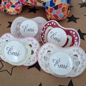 May include: Four pacifiers with pink and white accents. Each pacifier has a white disc with the name "Émi" written in black.