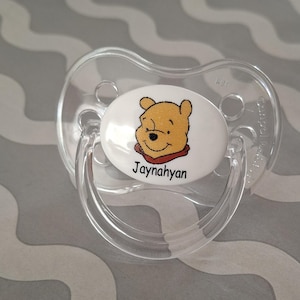 Peut inclure: Une sucette transparente pour bébé avec un motif Winnie l'ourson. La sucette présente un ovale blanc avec une image de dessin animé de Winnie l'ourson et le nom "Jaynahan" imprimé en noir. La sucette est en plastique transparent.