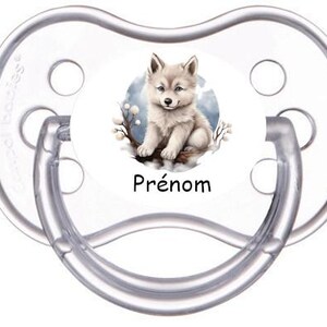 Peut inclure: Une sucette transparente pour bébé avec un motif rond blanc. Le motif représente une illustration de style aquarelle d'un louveteau duveteux et de couleur claire. Le mot "Prénom" est imprimé en noir en dessous.