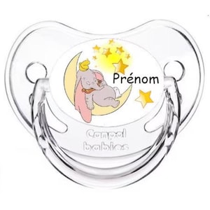 Peut inclure: Une sucette en silicone transparente avec un motif circulaire blanc représentant un éléphant de dessin animé dormant sur un croissant de lune jaune avec des étoiles jaunes. Le texte "Prénom" est imprimé sur le motif. La sucette est fabriquée par la marque "Compeed babies".