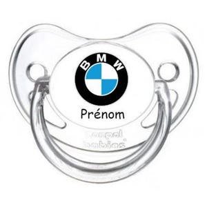 May include: A clear baby pacifier with a white center featuring the BMW logo in black, white, and blue. The word "Prénom" is printed below the logo. The pacifier is made of clear plastic.