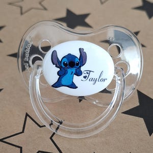 Peut inclure: Une sucette en plastique transparente avec un disque rond blanc représentant un personnage de dessin animé bleu avec le nom "Taylor" imprimé dessus.