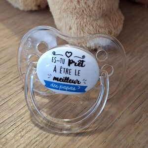 Peut inclure: Une sucette transparente avec un disque rond blanc et bleu qui dit "Es-tu prêt à être le meilleur des papas?" avec un symbole de cœur.
