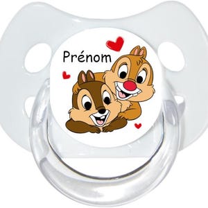 Könnte beinhalten: Ein klarer Babyschnuller mit einer weißen Scheibe mit Cartoon-Chipmunks und roten Herzen. Das Wort "Prénom" ist auf der Scheibe aufgedruckt. Der Schnuller hat einen klaren, abgerundeten Schild mit Luftlöchern.