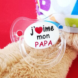 Peut inclure: Une sucette en plastique transparente avec un autocollant rond blanc qui dit "J'aime mon PAPA" avec un cœur rouge au milieu.