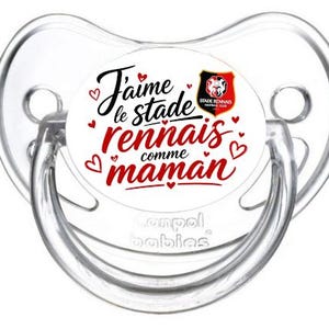 Peut inclure: Une sucette transparente pour bébé avec un motif circulaire blanc. Le motif affiche le texte "J'aime le stade rennais comme maman" en noir et rouge, avec des petits coeurs rouges et un logo d'équipe. La marque "Canpol babies" est également visible.