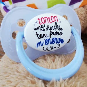 May include: A blue pacifier with a white oval-shaped tag that says "Tonton viens me chercher ton frère m'énerve".