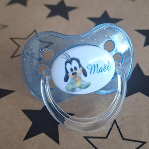 Peut inclure: Une sucette bleue avec un insert ovale blanc représentant un dessin animé d'un chien avec le nom "Maël" imprimé dessus.