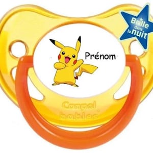 Peut inclure: Une sucette jaune et orange avec un autocollant blanc représentant un personnage de dessin animé Pikachu. L'autocollant comporte également le texte "Prénom" et "Compaal bébés". Un autocollant en forme d'étoile bleue avec le texte "Brille dans la nuit" se trouve dans le coin supérieur droit.