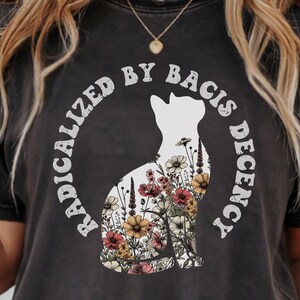 Könnte beinhalten: Dunkelgraues T-Shirt mit einer weißen Katzensilhouette, gefüllt mit bunten Blumen. Der Text "RADICALIZED BY BACIS DECENCY" ist kreisförmig um die Katze gedruckt. Das Shirt hat einen Vintage-Look.