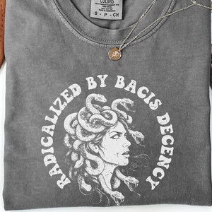 以下が含まれることがあります： メデューサのグラフィックを囲むように「RADICALIZED BY BACIS DECENCY」の文字がプリントされたグレーのComfort Colors Tシャツ。ペンダント付きのゴールドネックレスがシャツの上に置かれています。シャツは白い面に折りたたまれています。