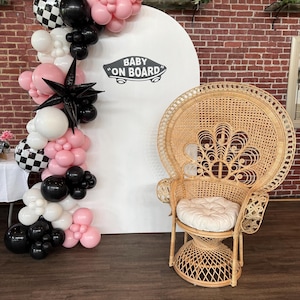 May include: A rattan peacock chair with a cream cushion sits next to a white backdrop with "Baby on Board" text. A balloon arch in black, white, and pink decorates the scene, suitable for a baby shower or nursery.