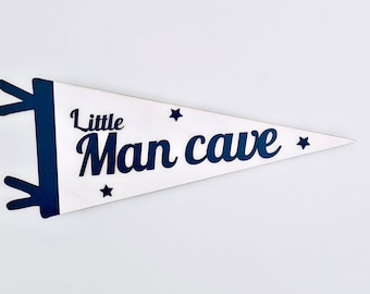 May include: A white pennant flag with navy blue accents and text that reads "Little Man cave". The flag has a triangular shape with navy blue decorative elements on the left side and small star shapes.