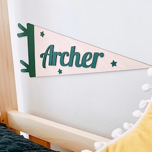 Peut inclure: Un fanion en bois avec le nom "Archer" en vert. Le fanion est de couleur claire avec des accents verts et des étoiles. Il est fixé sur un mur blanc près d'un cadre de lit en bois et d'un coussin jaune.