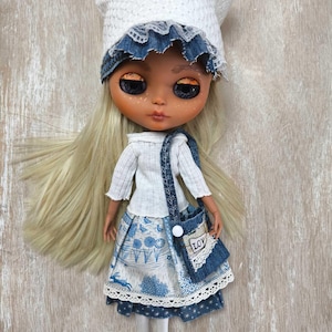May include: A doll with blonde hair wearing a white crocheted hat, a white sweater, a blue and white skirt with a floral pattern, and a blue denim bag with the word "Love" on it.