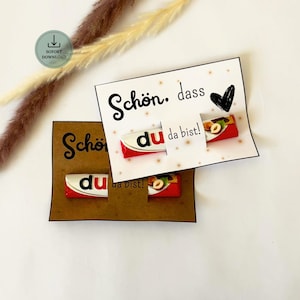 Puede incluir: Dos tarjetas rectangulares de papel, una blanca y otra marrón, cada una con una pequeña barra de chocolate. La tarjeta blanca presenta el texto "Schön, dass du da bist!" con un corazón negro. La tarjeta marrón tiene el mismo texto y una avellana.