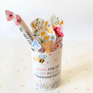 Puede incluir: Un recipiente cil&iacute;ndrico de papel decorado con ilustraciones de abejas y flores, con el texto en alem&aacute;n "DANKE F&Uuml;R DIE KUNTERBUNTE KINDERGARTENZEIT". Contiene recortes de papel de flores, un oso y una etiqueta con forma de mariposa con texto.