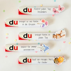 Puede incluir: Cinco barras de chocolate envueltas en fundas de papel blanco, cada una con la palabra du en negro y rojo, seguida de texto en alem&aacute;n e ilustraciones que incluyen mariposas rosas, flores coloridas, un conejo marr&oacute;n y una abeja amarilla.