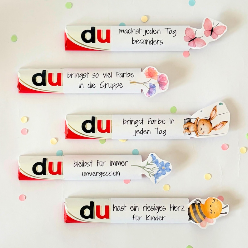 Puede incluir: Cinco barras de chocolate envueltas en fundas de papel blanco, cada una con la palabra du en negro y rojo, seguida de texto en alem&aacute;n e ilustraciones que incluyen mariposas rosas, flores coloridas, un conejo marr&oacute;n y una abeja amarilla.