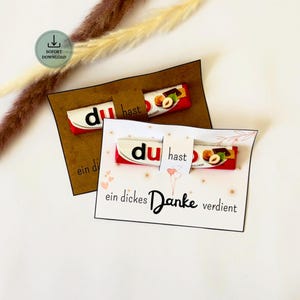 Może przedstawiać: Dwie prostokątne papierowe karty z batonami czekoladowymi Duplo. Jedna karta jest brązowa z czarnym tekstem, druga biała z różowymi sercami i napisem: du hast ein dickes Danke verdient. W lewym górnym rogu widoczna jest zielona ikona pobierania.