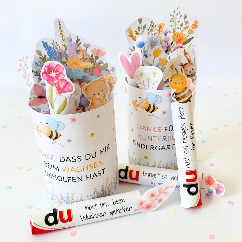 Puede incluir: Dos recipientes cil&iacute;ndricos de papel decorados con ilustraciones florales coloridas, abejas y un osito de peluche. El texto dice: DANKE, DASS DU MIR BEIM WACHSEN GEHOLFEN HAST, DANKE F&Uuml;R KUNTERBUNTE KINDERGARTEN, du hast ein riesiges Herz f&uuml;r Kinder, du bringst so viel in die Welt.