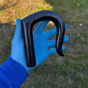 Puede incluir: Un mango de bastón negro y curvado. El mango lo sostiene una persona que lleva guantes azules y una sudadera azul. El mango tiene un acabado liso y pulido y un diseño curvado.