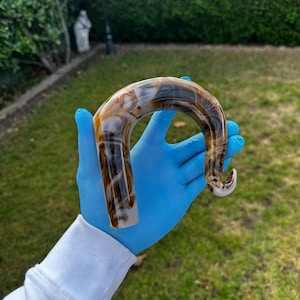 Puede incluir: Un mango de bastón curvo con un patrón veteado en tonos marrón, beige y blanco. El mango tiene un acabado liso y brillante y un diseño curvo. El mango lo sostiene una persona con guantes azules.