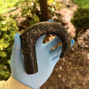May include: A black cane handle with a curved top and a straight shaft. The handle has a gold, speckled pattern. The cane handle is held by a person wearing a blue glove.
