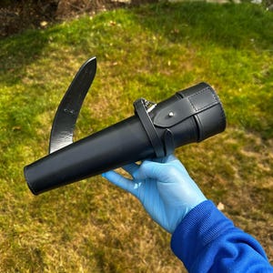 K&ouml;nnte beinhalten: Eine schwarze Lederscheide mit einer gebogenen Klinge und einem zylindrischen K&ouml;rper. Die Scheide hat einen Riemen mit einer Schnalle. Die Klinge ist etwa 30 cm lang. Der Hintergrund ist gr&uuml;nes Gras.