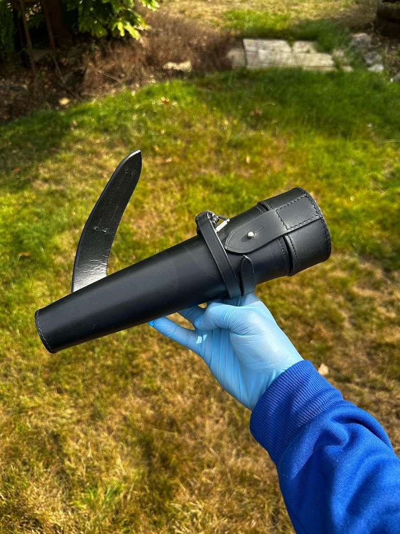 K&ouml;nnte beinhalten: Eine schwarze Lederscheide mit einer gebogenen Klinge und einem zylindrischen K&ouml;rper. Die Scheide hat einen Riemen mit einer Schnalle. Die Klinge ist etwa 30 cm lang. Der Hintergrund ist gr&uuml;nes Gras.