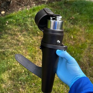 K&ouml;nnte beinhalten: Eine schwarze Lederflasche mit silbernem Deckel, umh&uuml;llt von einer passenden Lederhalterung mit Klappenverschluss. Die Halterung hat eine G&uuml;rtelschlaufe. Die Flasche wird im Freien vor einem gr&uuml;nen, grasbewachsenen Hintergrund gezeigt.