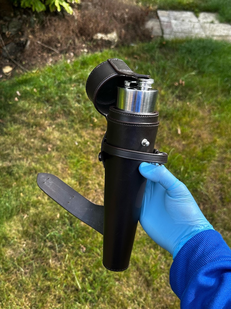 K&ouml;nnte beinhalten: Eine schwarze Lederflasche mit silbernem Deckel, umh&uuml;llt von einer passenden Lederhalterung mit Klappenverschluss. Die Halterung hat eine G&uuml;rtelschlaufe. Die Flasche wird im Freien vor einem gr&uuml;nen, grasbewachsenen Hintergrund gezeigt.