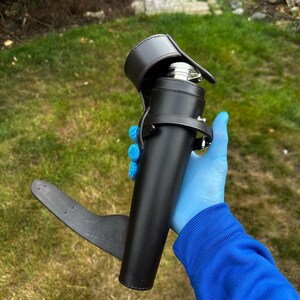 K&ouml;nnte beinhalten: Ein schwarzer Lederflaschenhalter mit passendem Riemen und einer silbernen Flasche im Inneren. Der Halter hat eine Klappe, die die Oberseite der Flasche bedeckt. Der Halter wird vor einem gr&uuml;nen, grasbewachsenen Hintergrund gehalten.