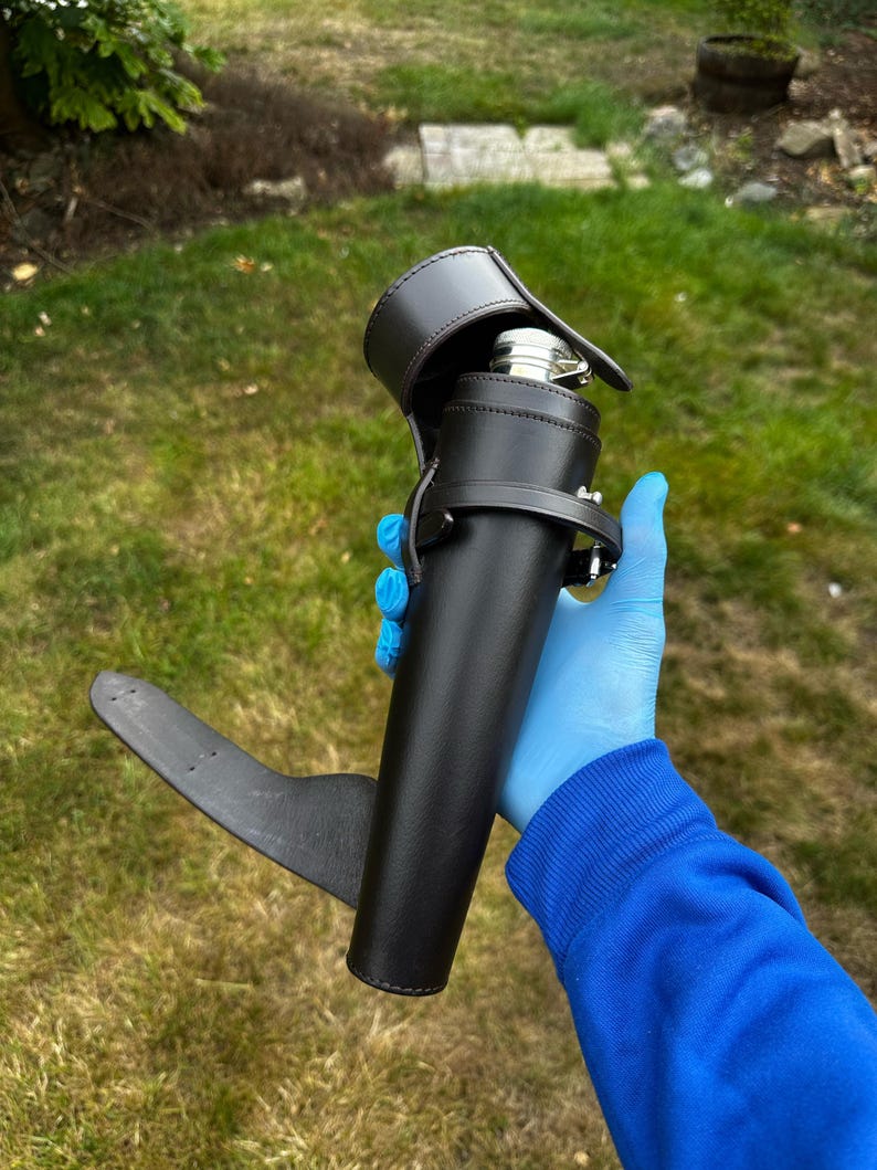 K&ouml;nnte beinhalten: Ein schwarzer Lederflaschenhalter mit passendem Riemen und einer silbernen Flasche im Inneren. Der Halter hat eine Klappe, die die Oberseite der Flasche bedeckt. Der Halter wird vor einem gr&uuml;nen, grasbewachsenen Hintergrund gehalten.