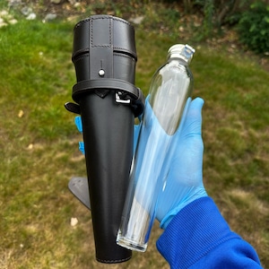 K&ouml;nnte beinhalten: Ein schwarzer Leder-Flachmannhalter mit silberner Schnalle und einer durchsichtigen Glasflasche. Die Flasche ist zylindrisch mit einem silbernen Verschluss. Der Halter hat einen G&uuml;rtel und einen Riemen. Der Hintergrund ist gr&uuml;nes Gras und Laub.