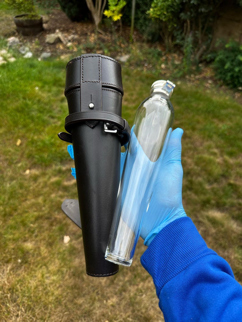 K&ouml;nnte beinhalten: Ein schwarzer Leder-Flachmannhalter mit silberner Schnalle und einer durchsichtigen Glasflasche. Die Flasche ist zylindrisch mit einem silbernen Verschluss. Der Halter hat einen G&uuml;rtel und einen Riemen. Der Hintergrund ist gr&uuml;nes Gras und Laub.