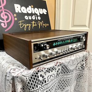Puede incluir: Un receptor AM/FM de estado sólido Sanyo vintage con acabado de veta de madera. La placa frontal es plateada con múltiples perillas y diales. El texto "Radique audio Enjoy the Music" está en el fondo.