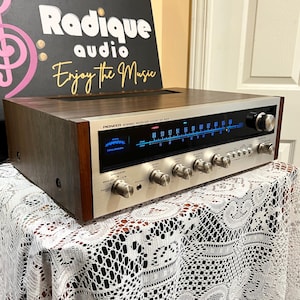 Puede incluir: Un receptor estéreo Pioneer SX-525 vintage con acabado de veta de madera. La placa frontal es plateada con múltiples perillas y diales. La pantalla muestra un dial azul iluminado. El texto "Radique audio Enjoy the Music" está en el fondo.