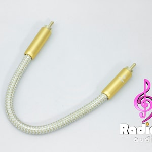 Puede incluir: Un cable de audio de color dorado con una capa exterior trenzada blanca. El cable tiene dos conectores RCA en cada extremo.