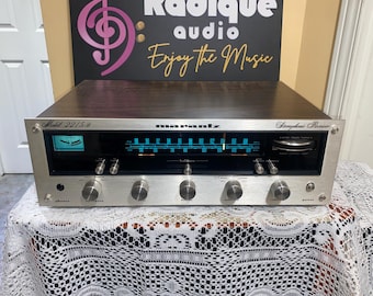 Może przedstawiać: Srebrny odbiornik stereo Marantz 2215B z ciemnym drewnianym blatem. Przód ma wiele pokręteł, tarcz i niebieski podświetlany wyświetlacz. Widoczny jest napis "Symphonic Receiver".