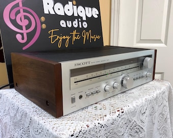 Puede incluir: Un receptor estéreo Scott vintage con una cara plateada, paneles laterales de madera y una parte superior negra. El receptor tiene múltiples perillas y diales. El texto "Radique audio Enjoy the Music" es visible en el fondo.