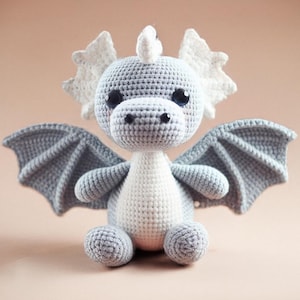 May include: A handmade crochet dragon toy in shades of gray and white. The dragon has large, round eyes, a small snout, and detailed wings. The wings and head accents are a lighter shade of gray, while the body is primarily white.