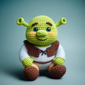 Pode incluir: Boneca de crochê feita à mão de um ogro verde com orelhas grandes, olhos azuis e uma expressão sorridente. A boneca usa uma camisa branca com um colete marrom e sapatos marrons. A boneca está sentada.