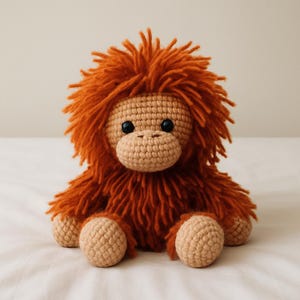 Könnte beinhalten: Ein handgefertigtes Orang-Utan-Plüschtier aus Häkelarbeit. Das Spielzeug hat ein beiges Gesicht und Gliedmaßen mit einem zotteligen, orangebraunen Fellkörper und Kopf. Es hat schwarze Knopfaugen und eine beige, strukturierte Schnauze. Das Plüschtier sitzt auf einer weißen Oberfläche.