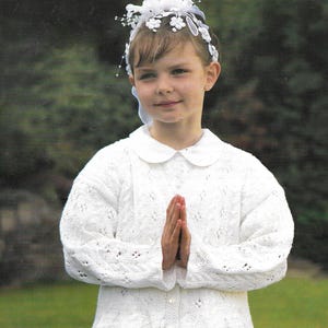 May include: A young girl wearing a white crocheted cardigan with a white collar and a white flower crown. She is standing in front of a green background and has her hands clasped in front of her.